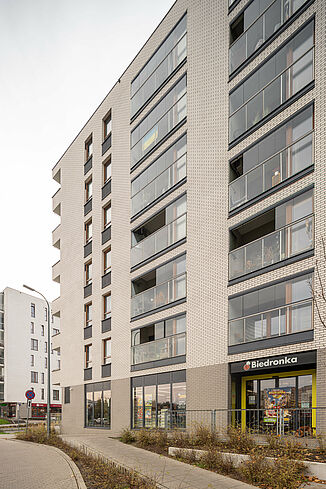 Widok z boku budynku w Warszawie z supermarketem na parterze i mieszkaniami powyżej.