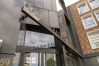 Wejście do nowoczesnego budynku z czarną fasadą, w którym mieści się szkoła muzyczna Léo Délibes w Paryżu.